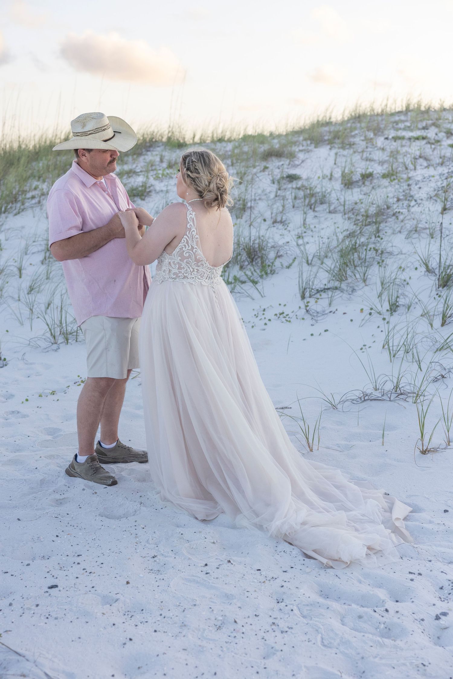 A couple shares a romantic moment on white sand dunes at sunset, with the bride wearing a flowing lace wedding gown.