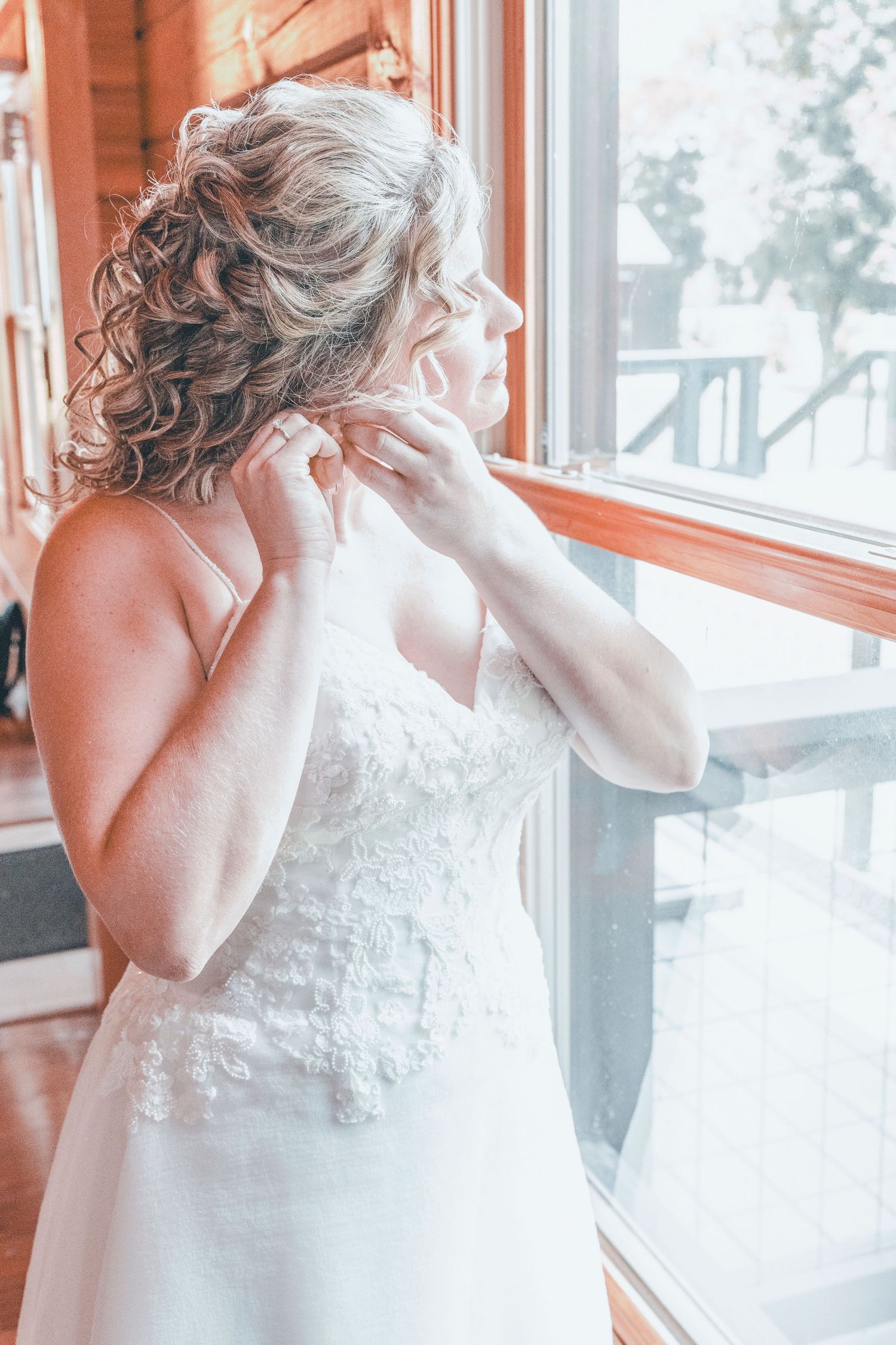 A person in a white lace wedding dress puts on earrings while standing by a window in soft natural light.