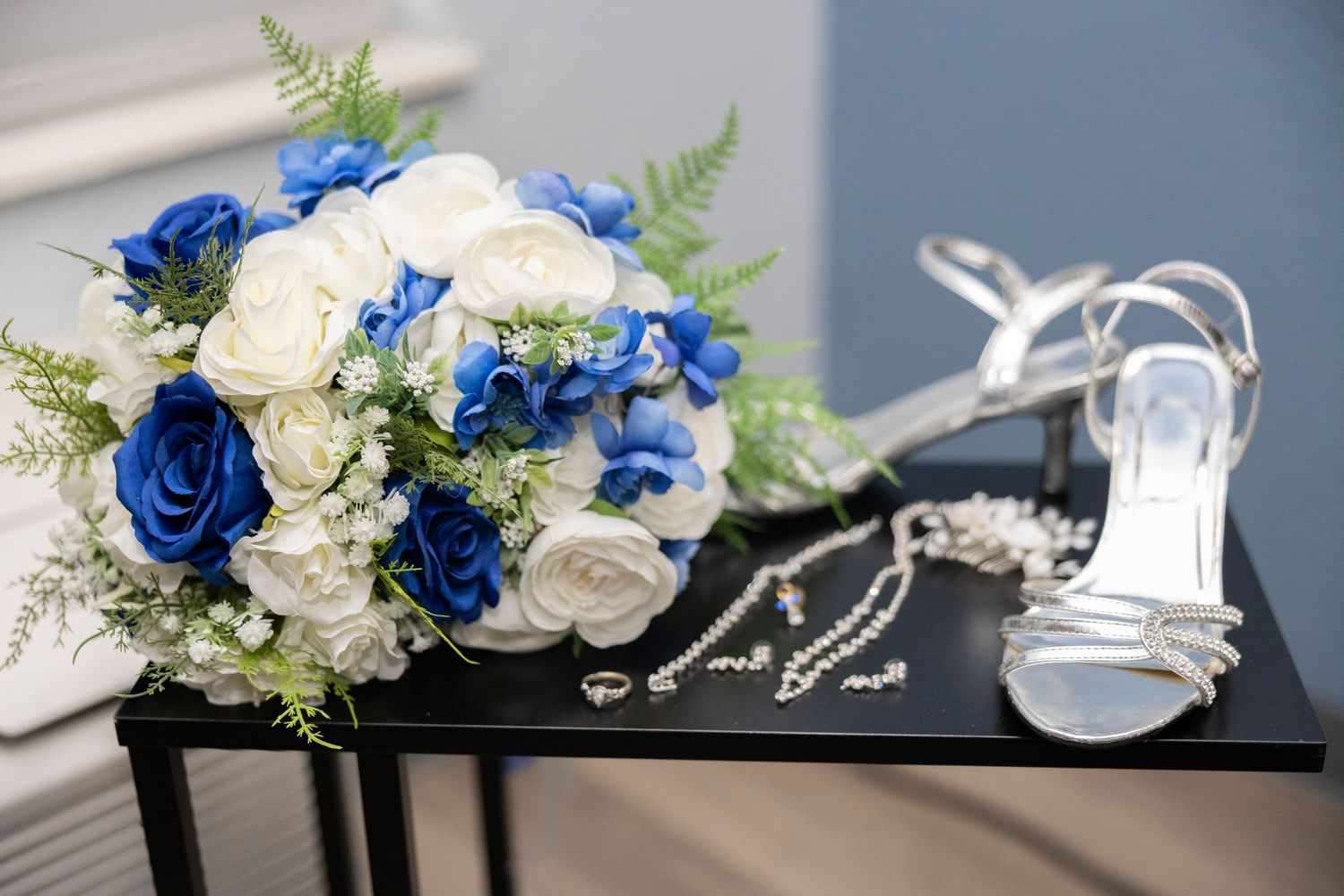 Wedding accessories including a blue and white rose bouquet, silver high heel sandals, and delicate jewelry on black surface.