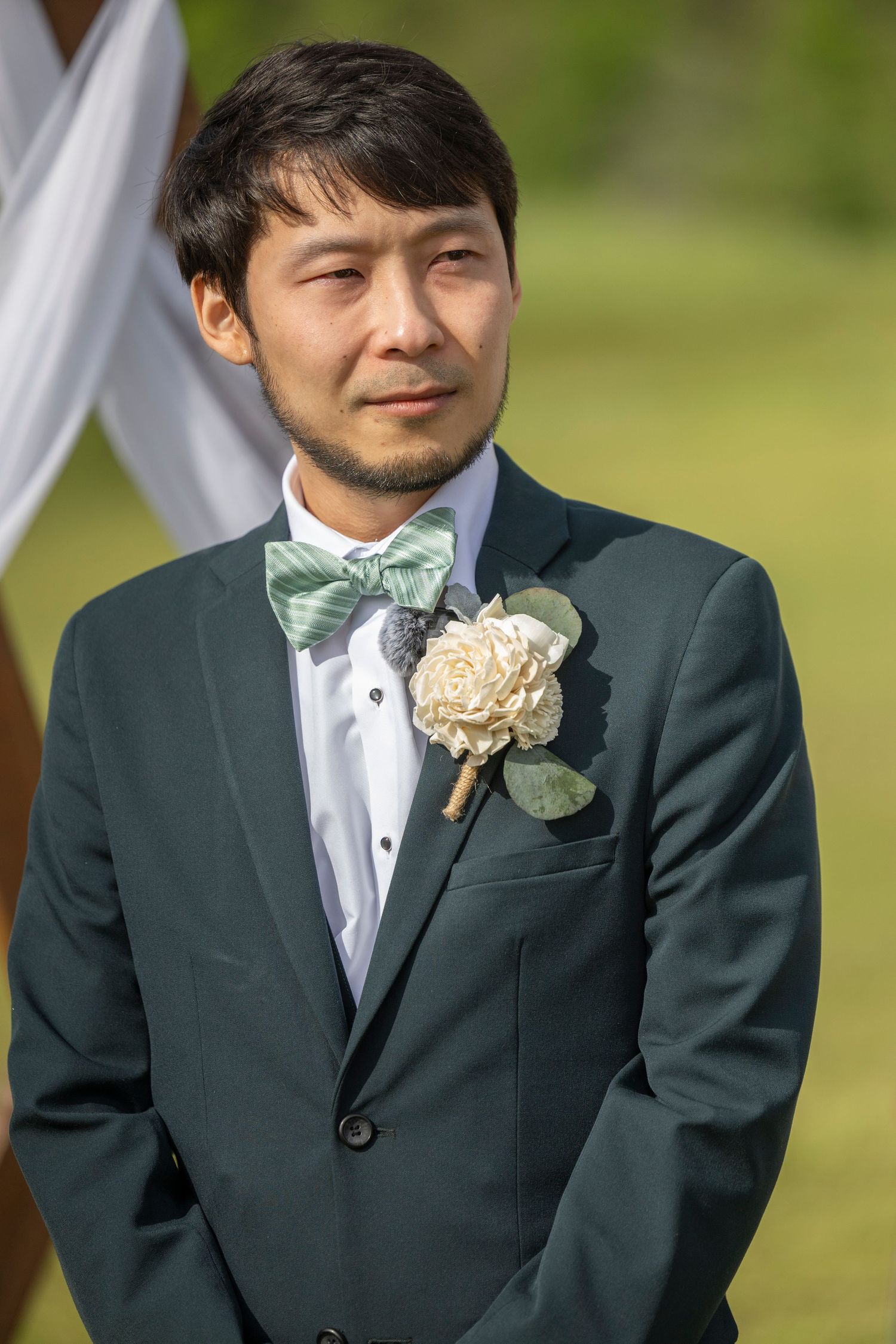 A groomsman in a dark green suit with a white boutonniere and bow tie smiles during an outdoor wedding ceremony.