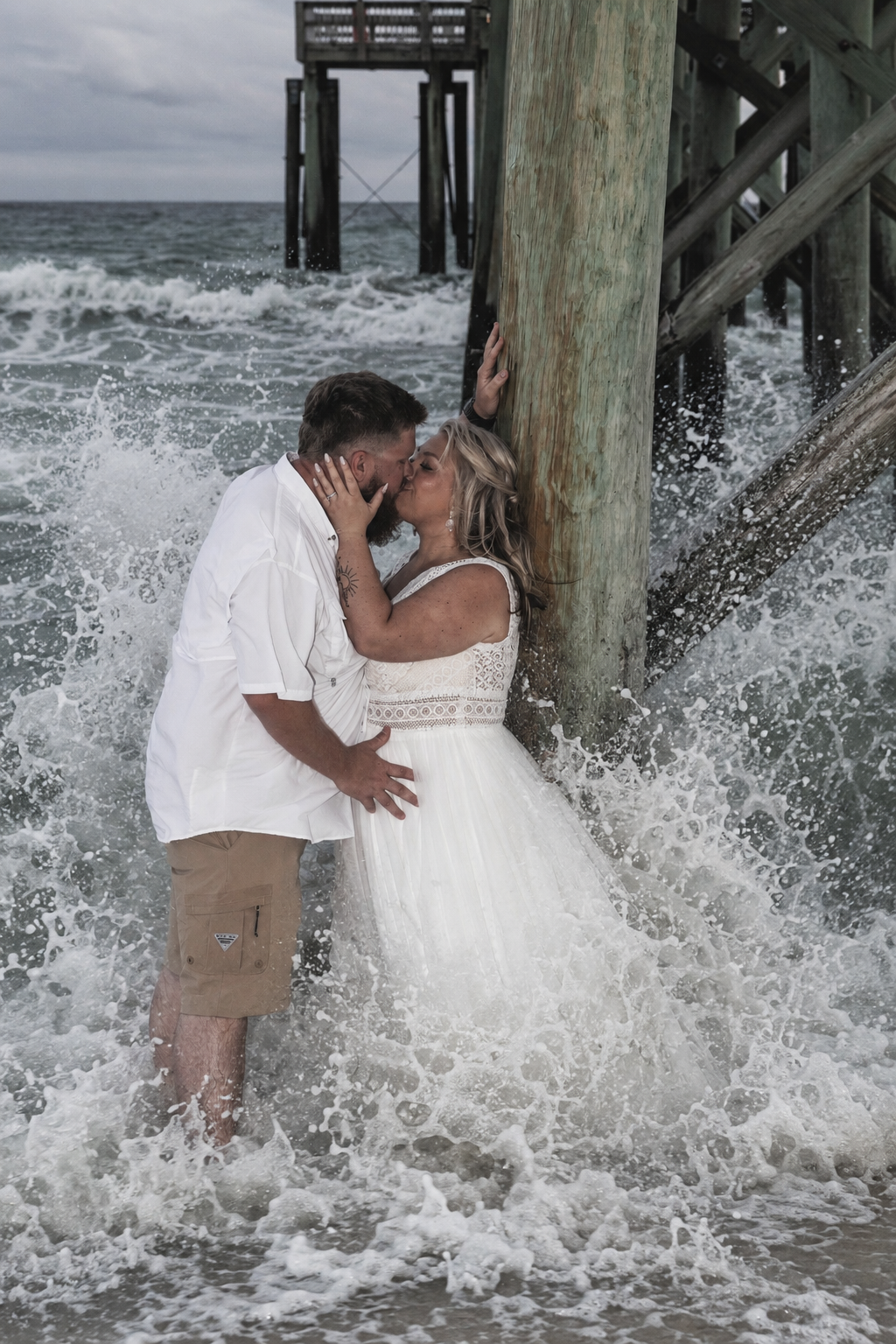 Couple kissing in ocean waves by pier, bride in white dress, groom in white shirt and khaki shorts.