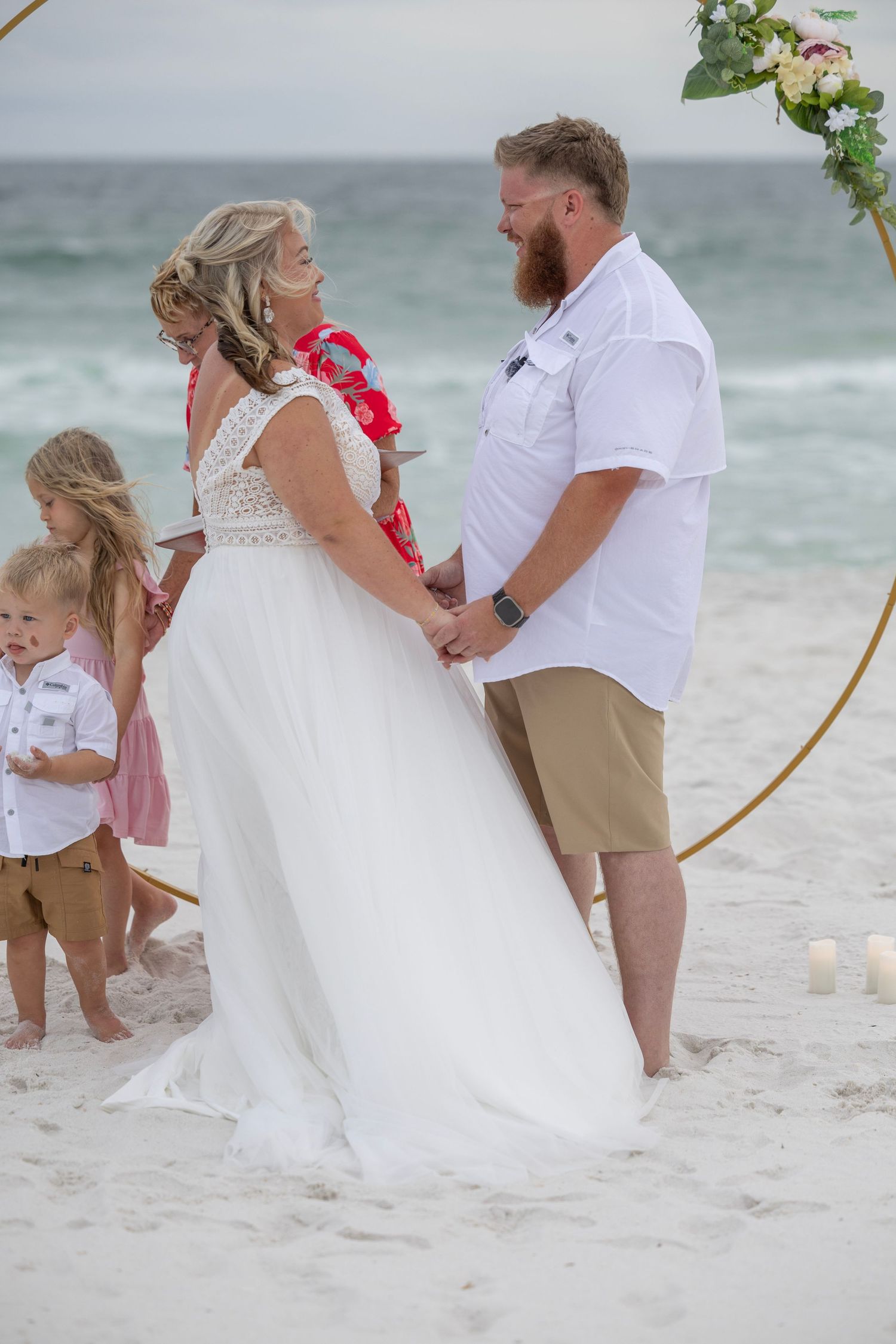 Couple exchanging vows at a romantic beach wedding ceremony under a floral arch with children nearby.