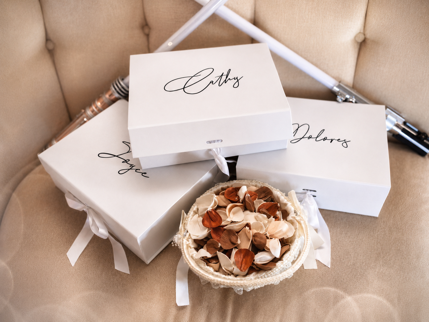 White personalized gift boxes with cursive names and ribbon bows, displayed with a small basket of decorative shells on a beige chair.