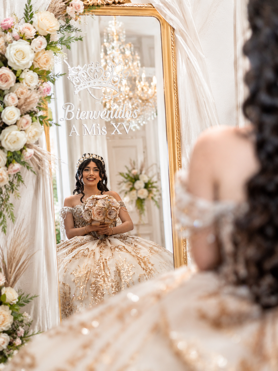 Young woman in gold quinceañera ball gown reflected in ornate mirror decorated with text Bienvenidos A Mis XV.
