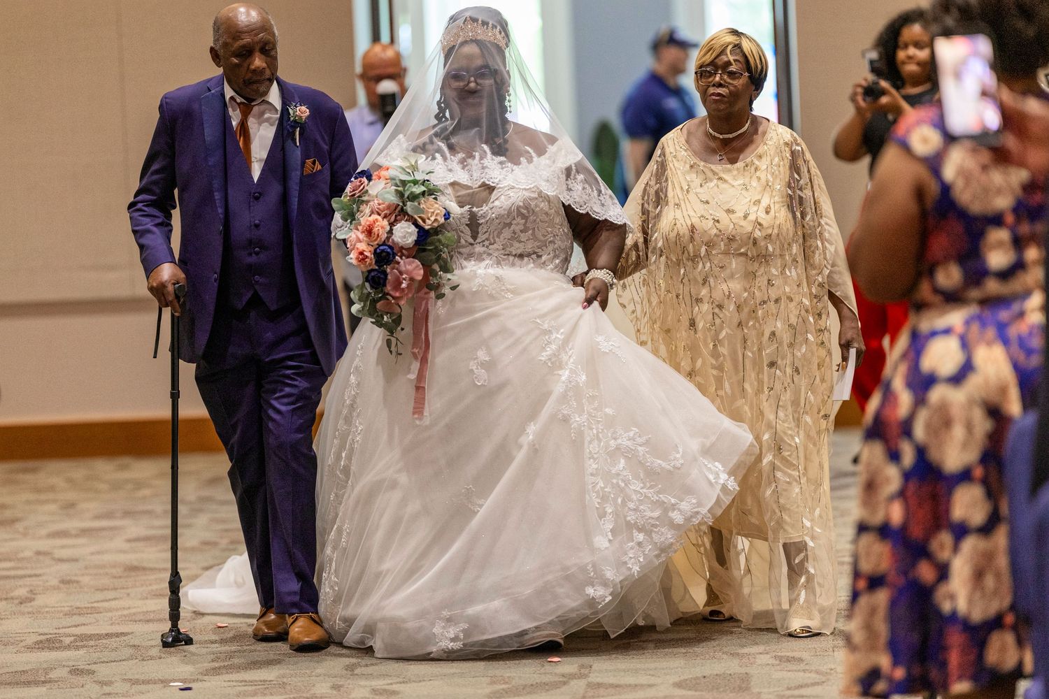 Bride in white gown walks down aisle escorted by elderly man with cane and woman in gold dress.