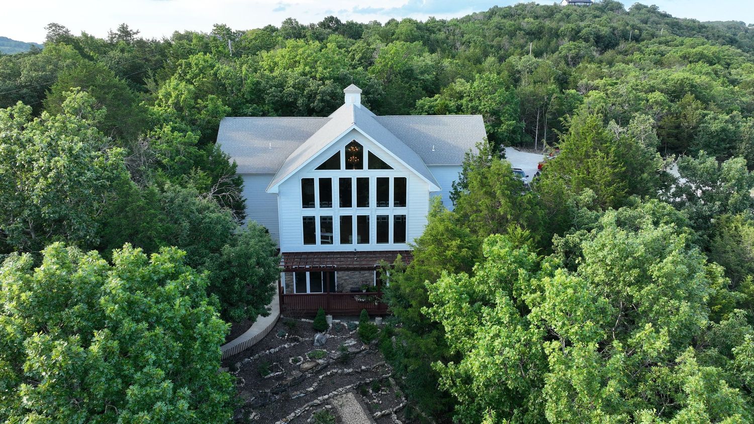 Aerial view of a large white A-frame house surrounded by lush green forest trees on a wooded hillside.