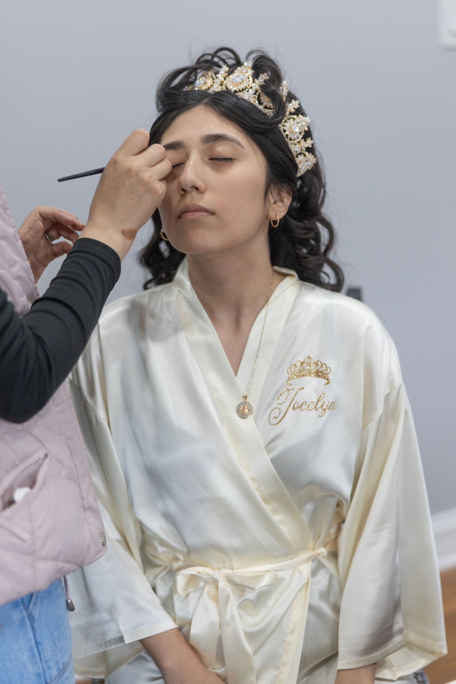 Bride named Jocelyn in white satin robe and gold crown gets makeup applied by artist before wedding.