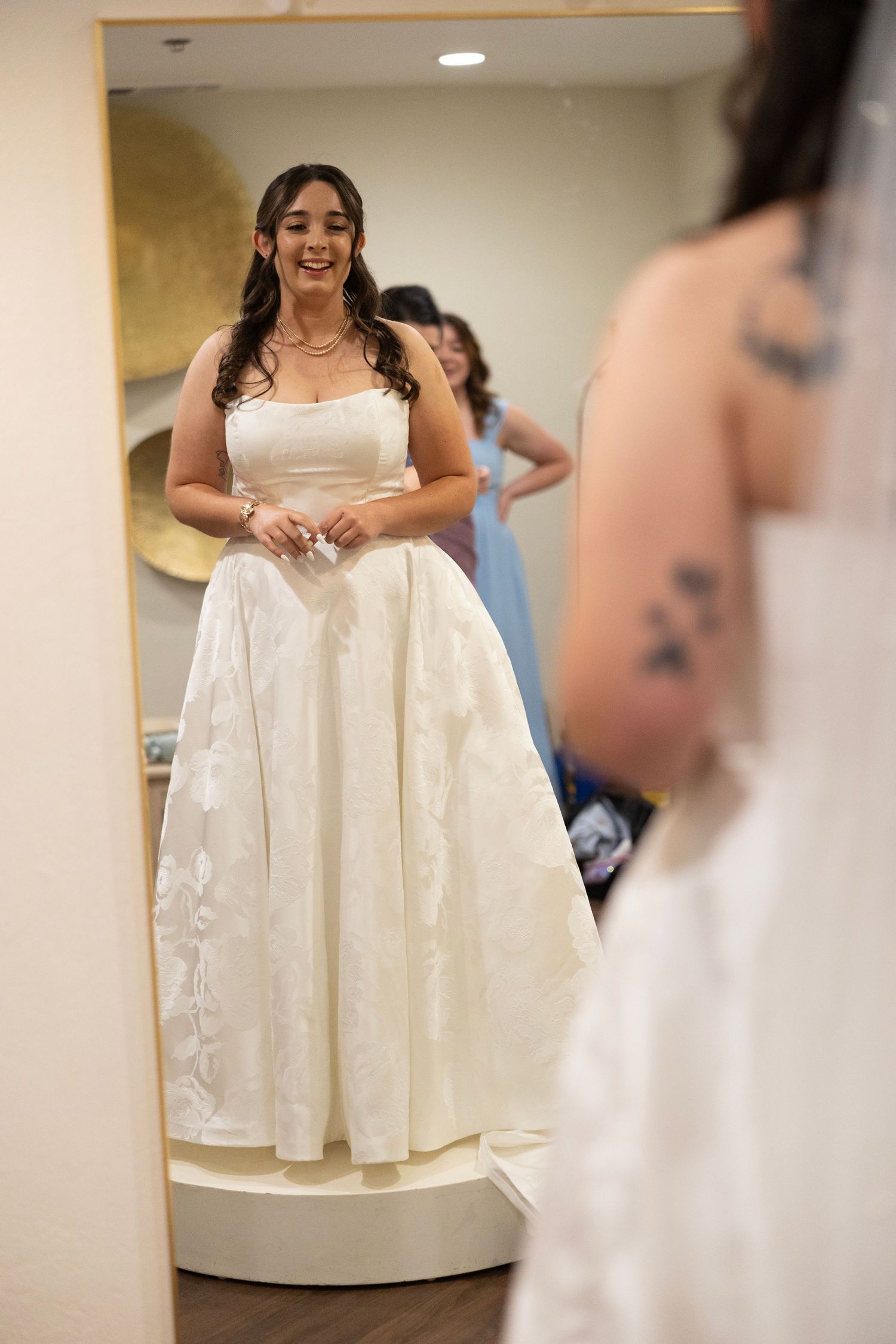 Bride smiling in a strapless white ball gown wedding dress while trying it on in a bridal boutique fitting room.