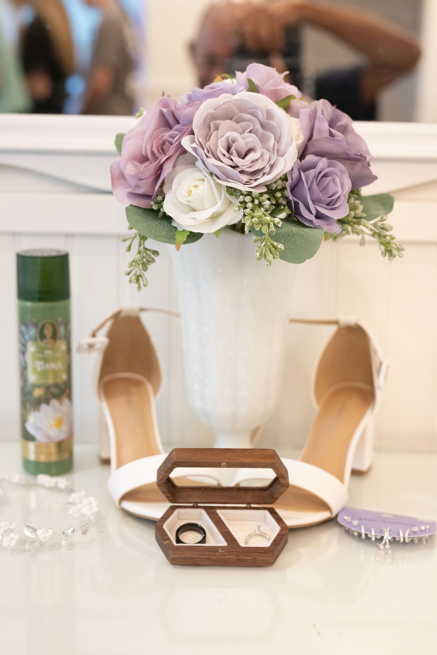 Bridal flat lay with purple roses bouquet, white heels, wooden ring box with wedding rings, and green hairspray on white surface.