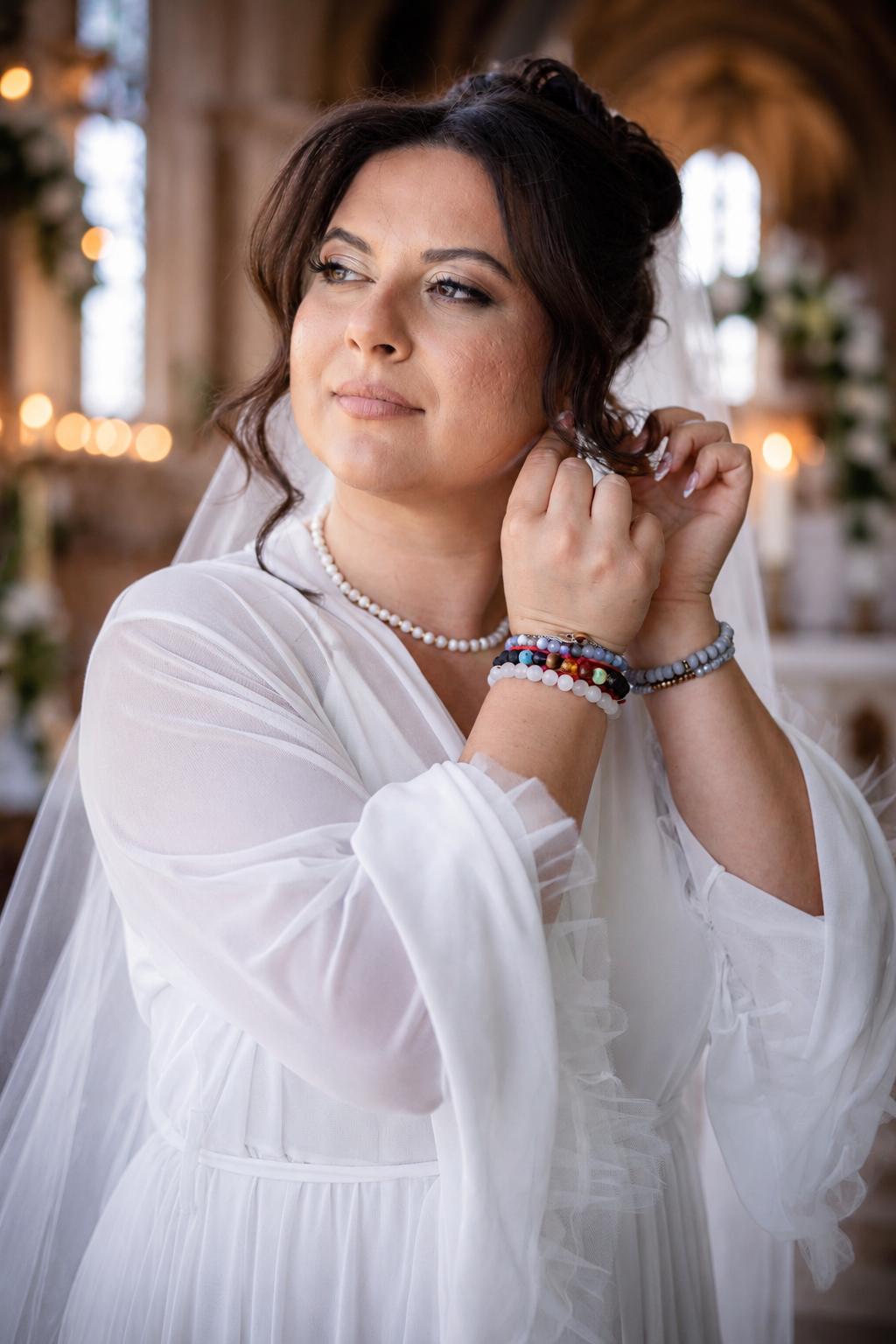 Bride in white sheer gown adjusting pearl earring, wearing colorful beaded bracelets, in elegant church setting.