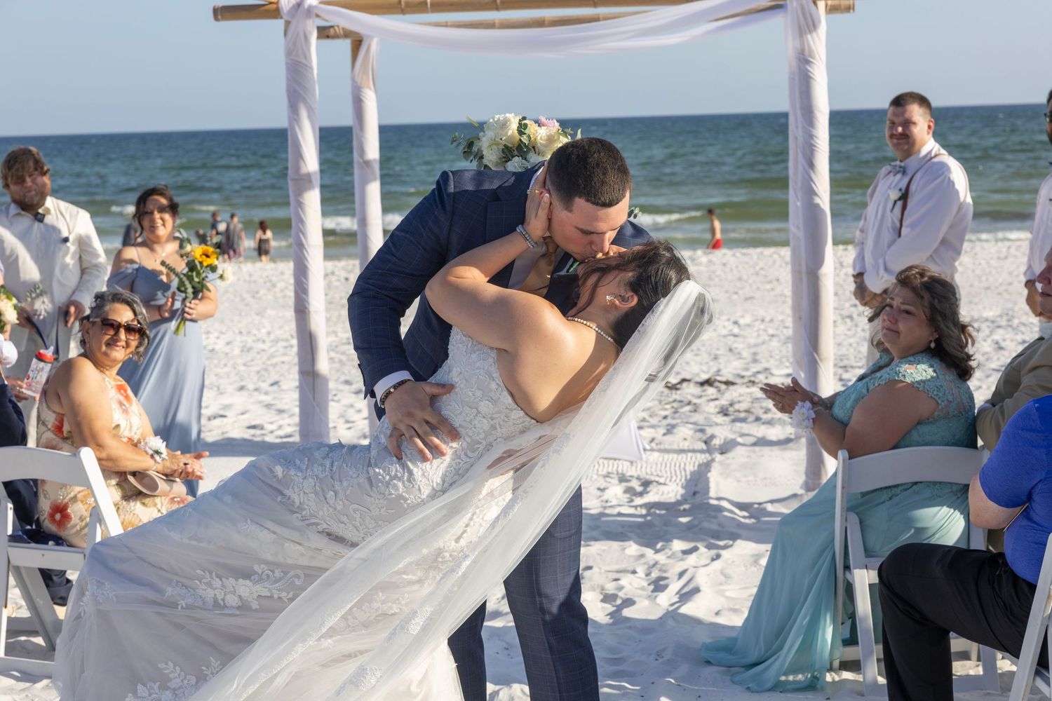 Bride and groom share romantic dip kiss at outdoor beach wedding ceremony with ocean backdrop and seated guests.