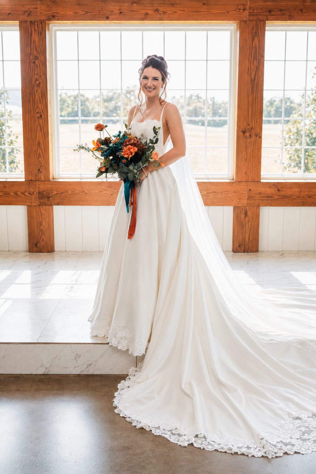 Elegant bride in a white A-line gown with lace trim holding a teal and orange bouquet inside a rustic barn venue.