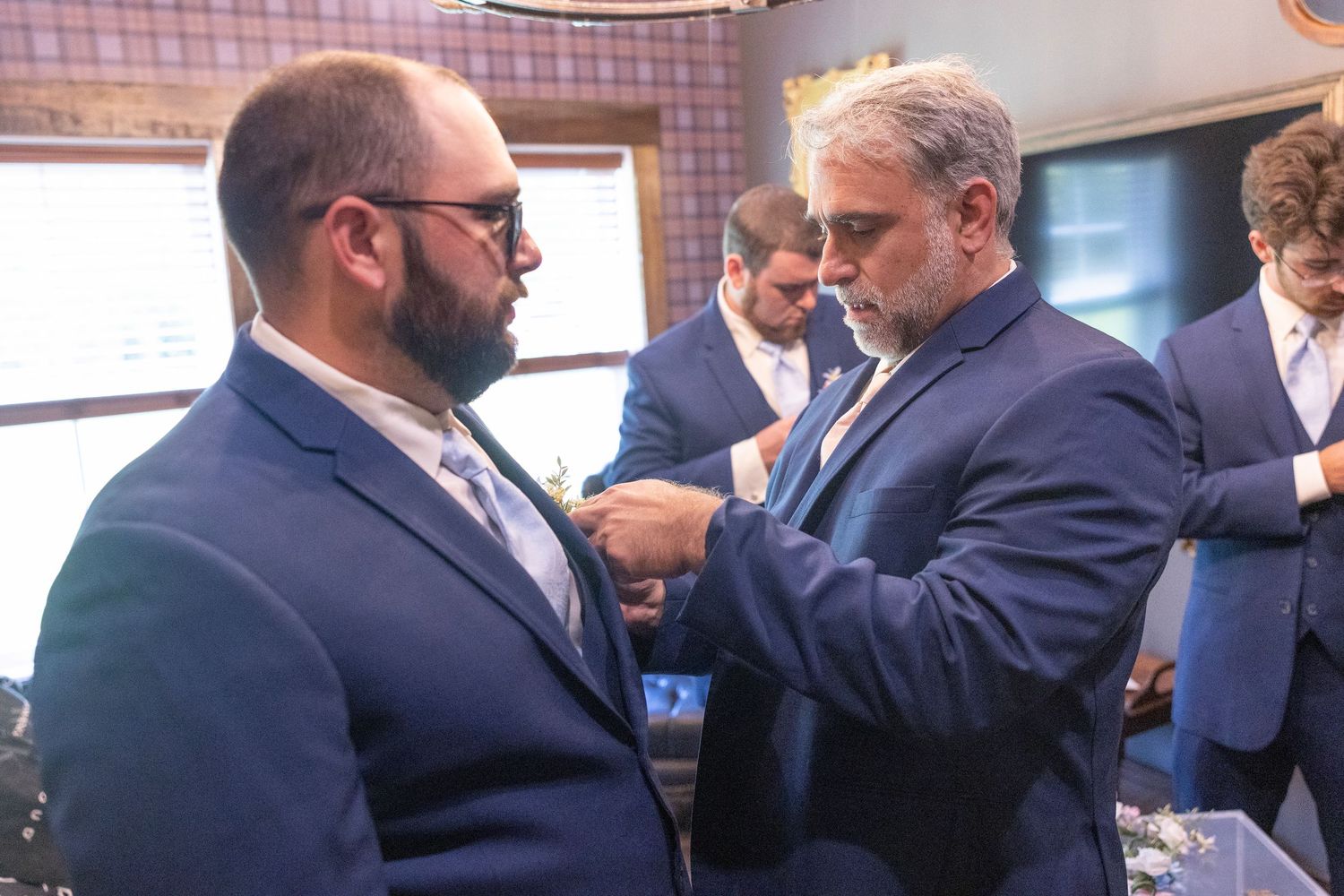 Two men in blue suits adjusting a tie or boutonniere before a formal event.