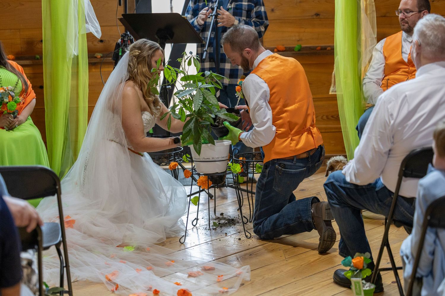 Bride and groom in orange vest perform unity plant ceremony indoors at rustic wedding venue with green decor.