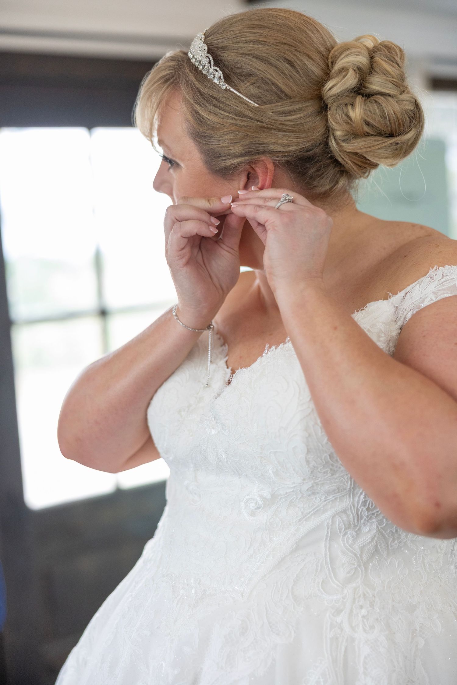 Bride in white lace gown and tiara puts on earrings while getting ready for her wedding day.