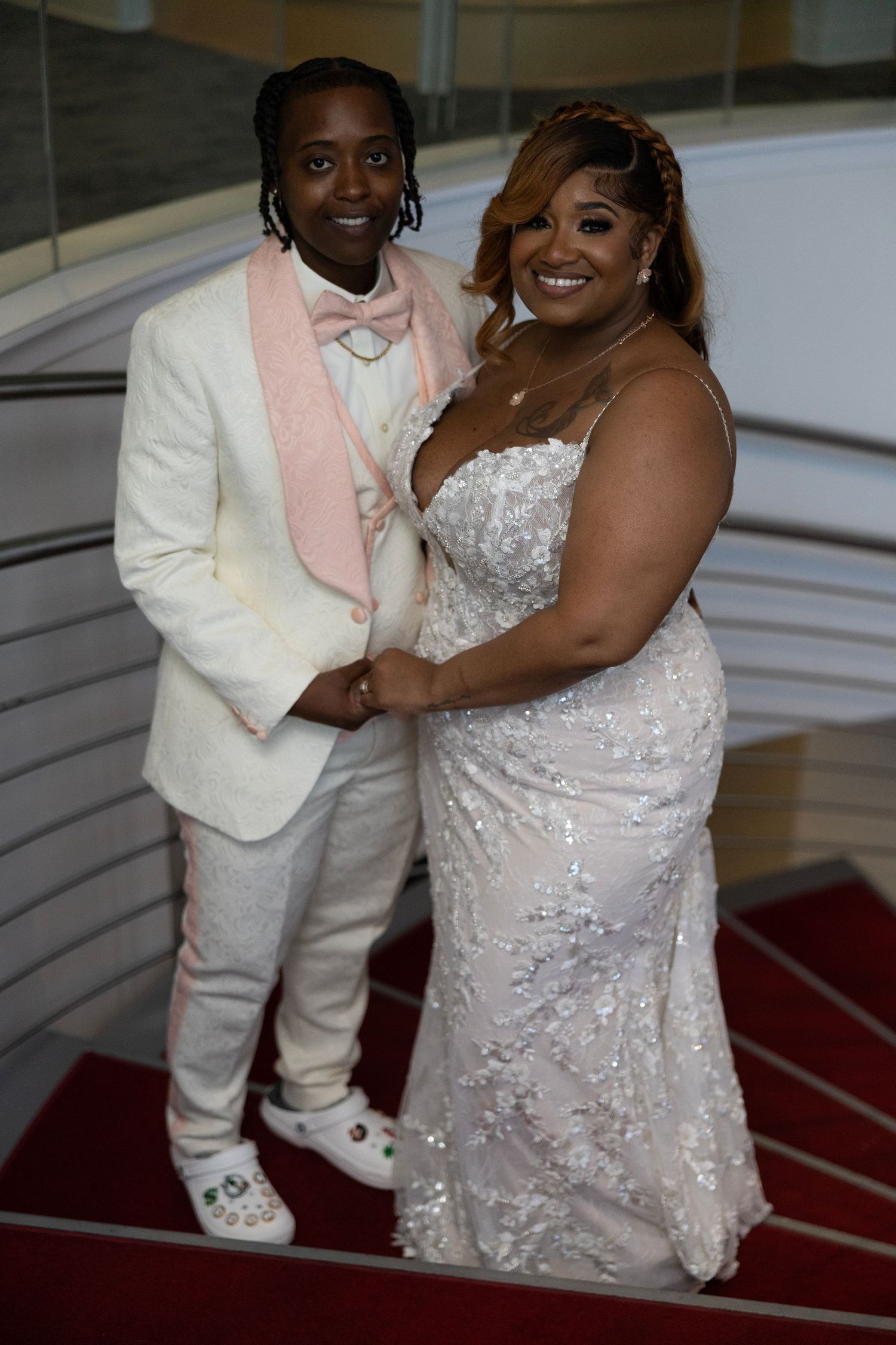 Couple posing on stairs in formal attire, man in white tuxedo with pink bow tie, woman in sequined white gown.