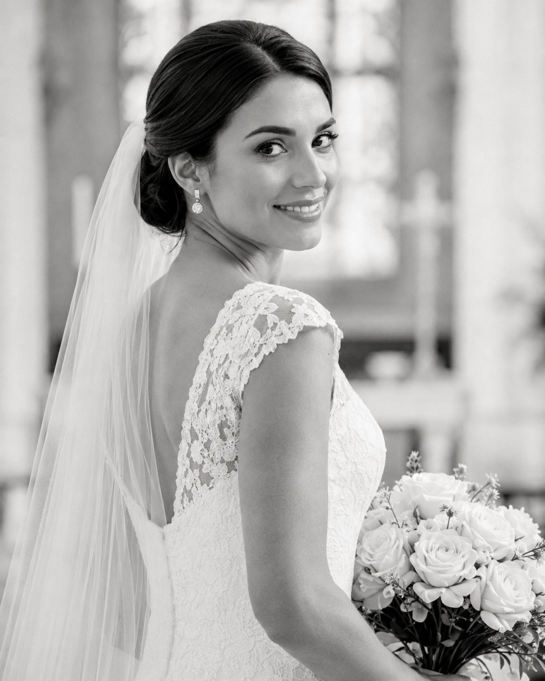 Black and white portrait of bride in lace gown holding bouquet with veil in church.