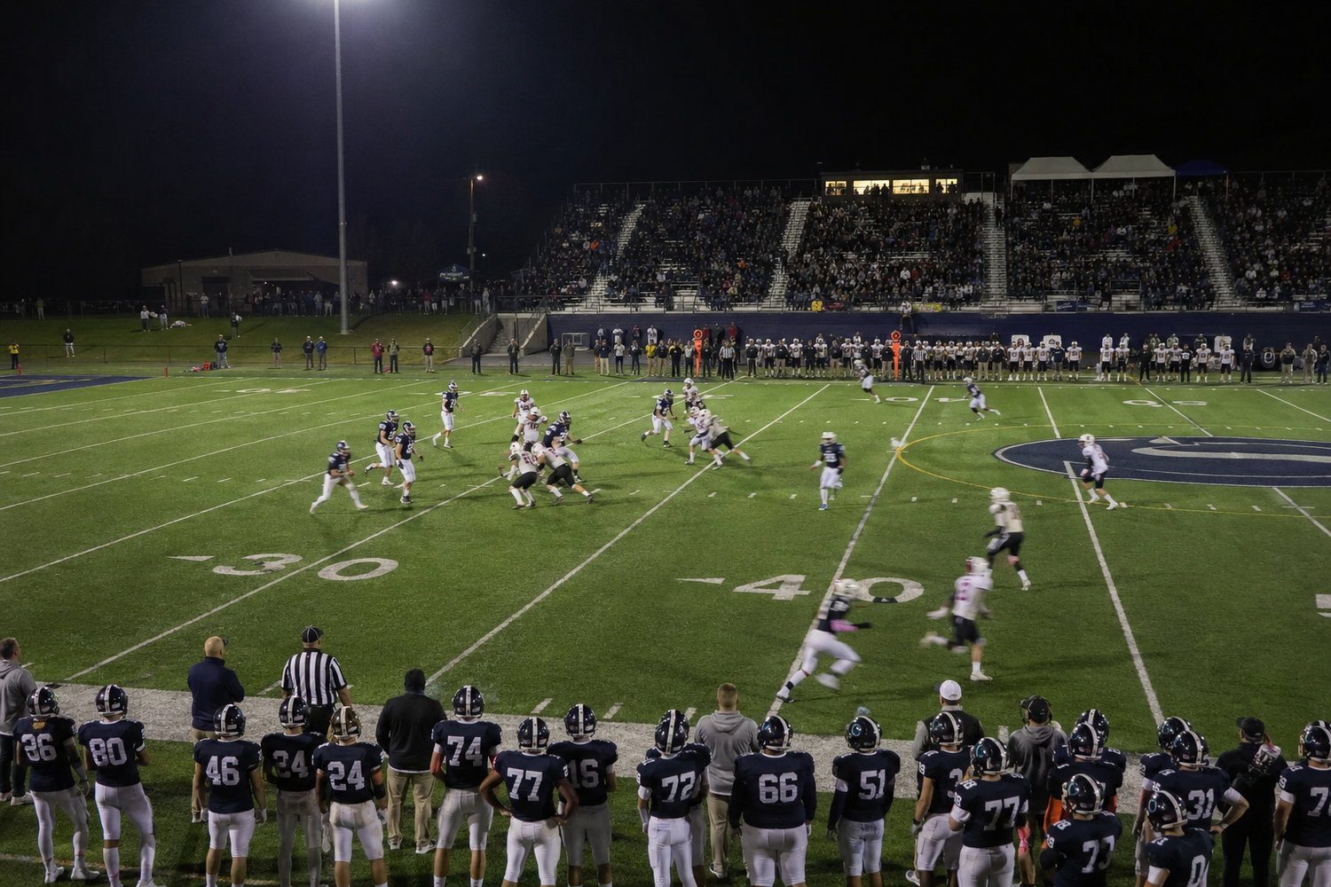 Wide-angle view of a night football game with players on the 40-yard line and coaches watching from the sideline.