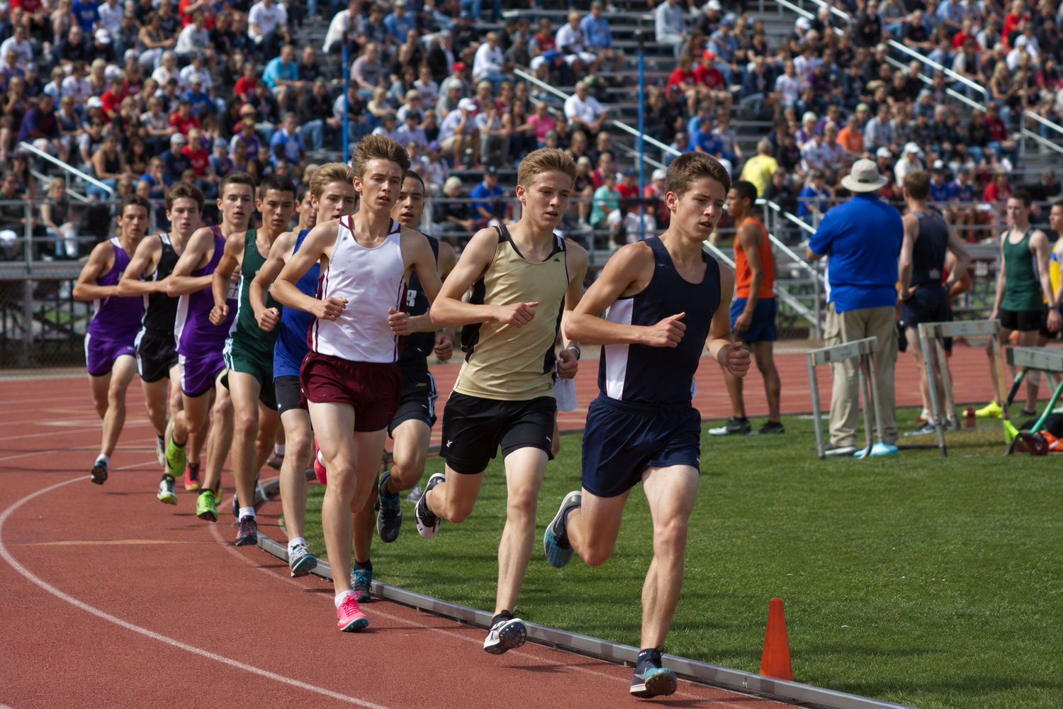 Runners compete in a high school track meet distance race on a red oval track with spectators in the stadium bleachers.
