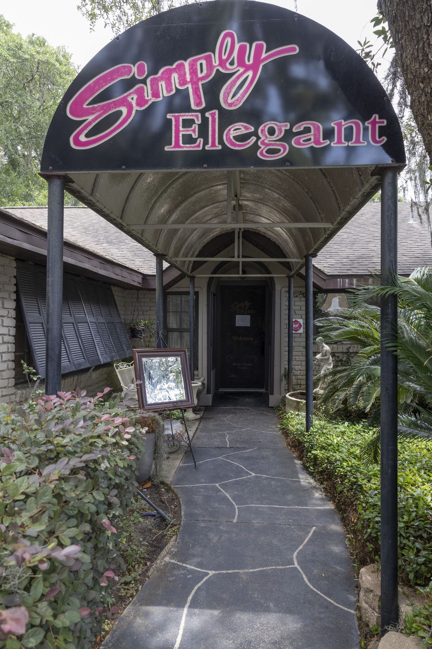 An entrance to 'Simply Elegant' business with a curved black awning and neon pink signage over a stone walkway.