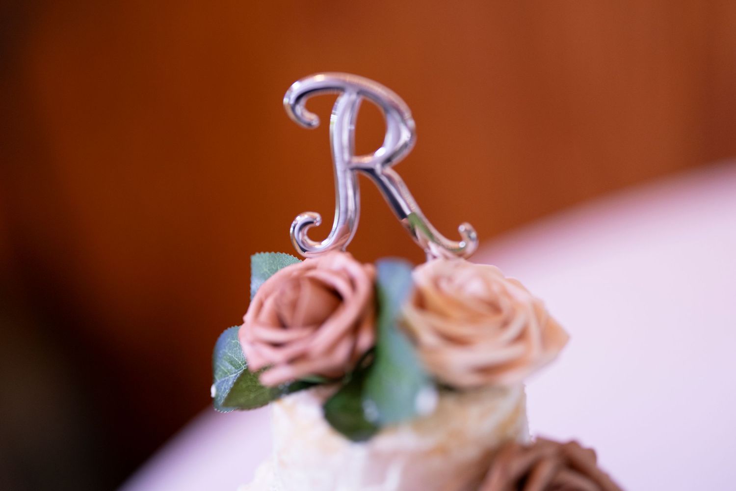 Ornate silver letter 'R' cake topper with blush pink roses in soft focus.
