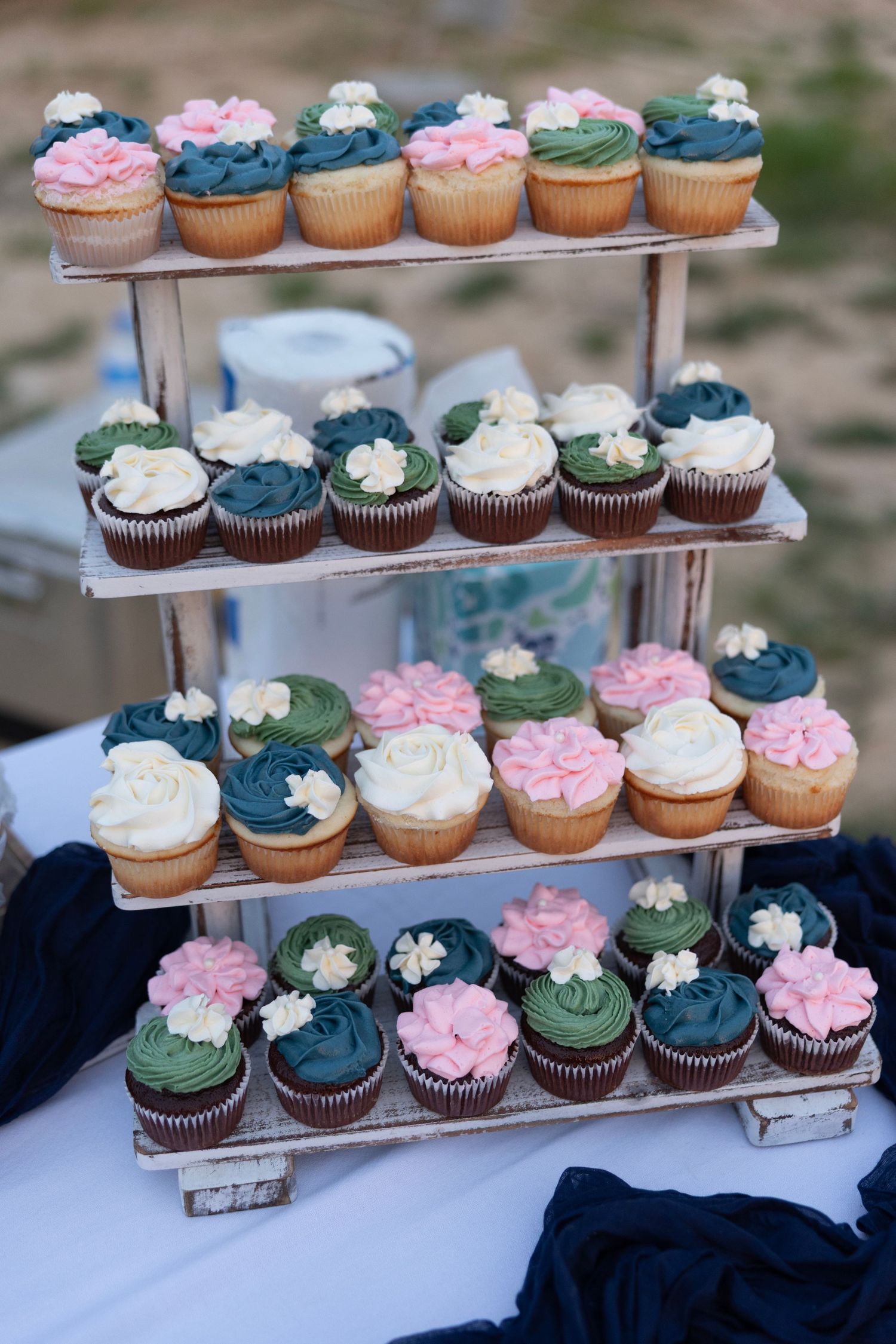 Four-tier rustic wooden stand displaying colorful cupcakes with pink, white, green, and navy blue frosting at a wedding.