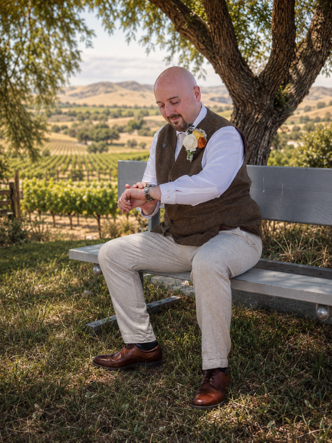 Groom in brown vest checks watch on bench at vineyard wedding venue with rolling hills backdrop.