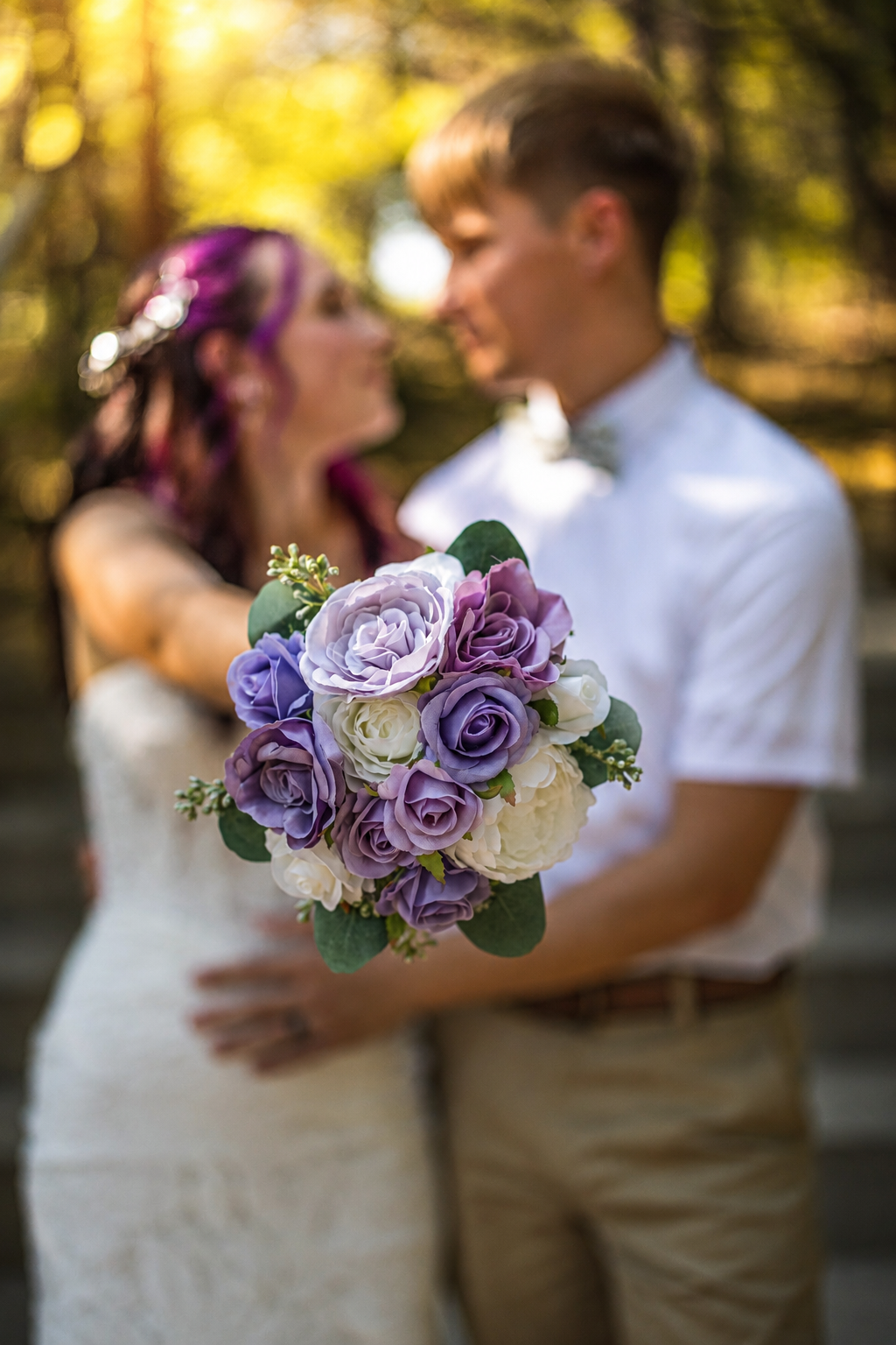 Wedding couple embracing outdoors while bride holds purple and white rose bouquet in soft bokeh light.