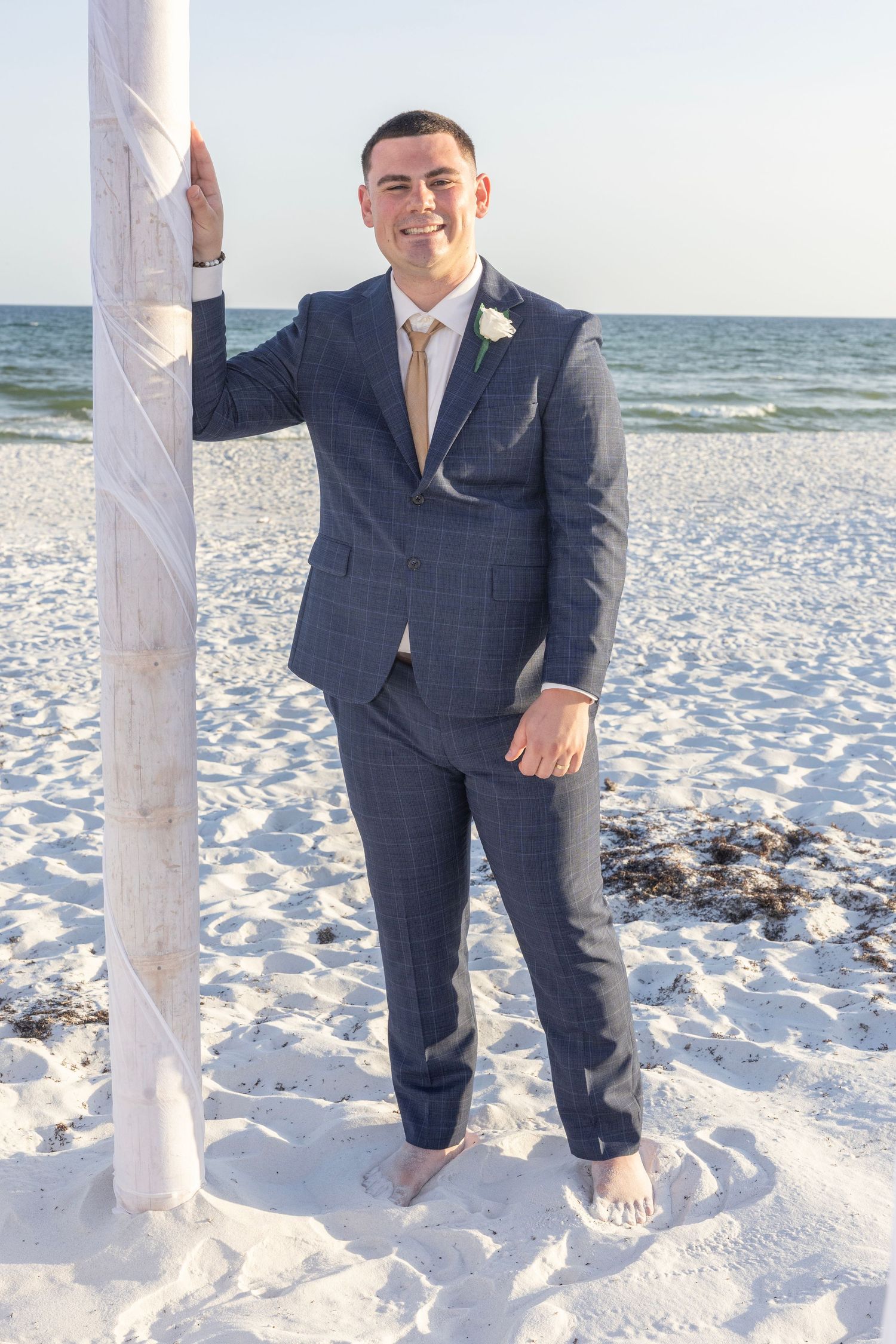 Groom in navy suit with white boutonniere stands barefoot on white sand beach, leaning against a pole with ocean in background.