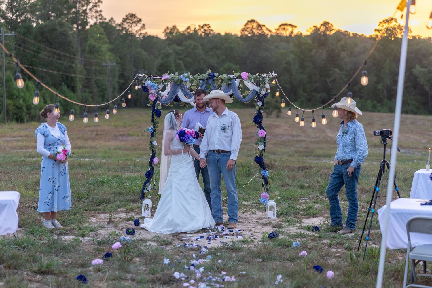Outdoor rustic wedding ceremony at sunset with bride and groom under floral arch, string lights, and guests in cowboy hats.