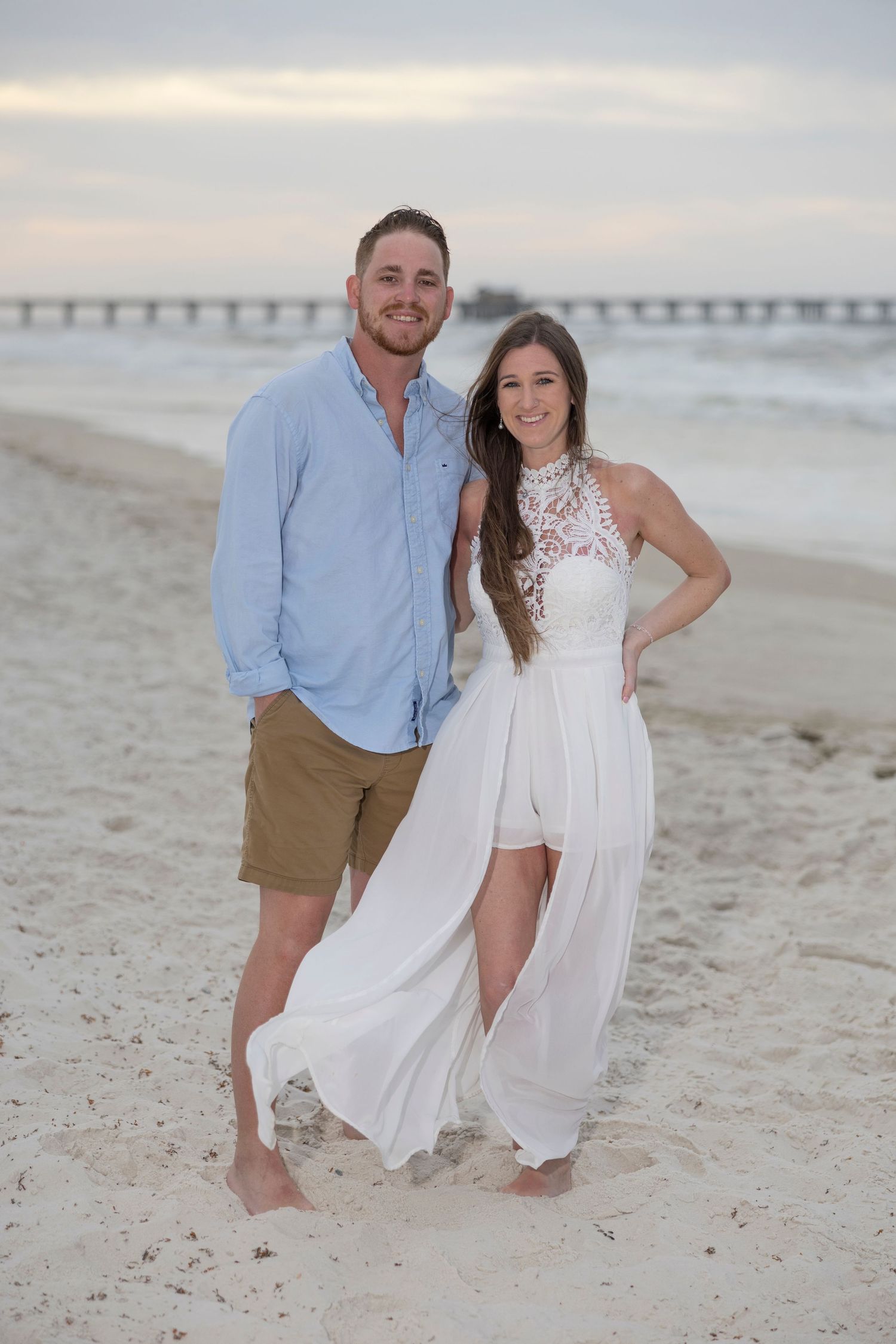 Couple posing on sandy beach at sunset, man in blue shirt and khaki shorts, woman in elegant white halter dress.