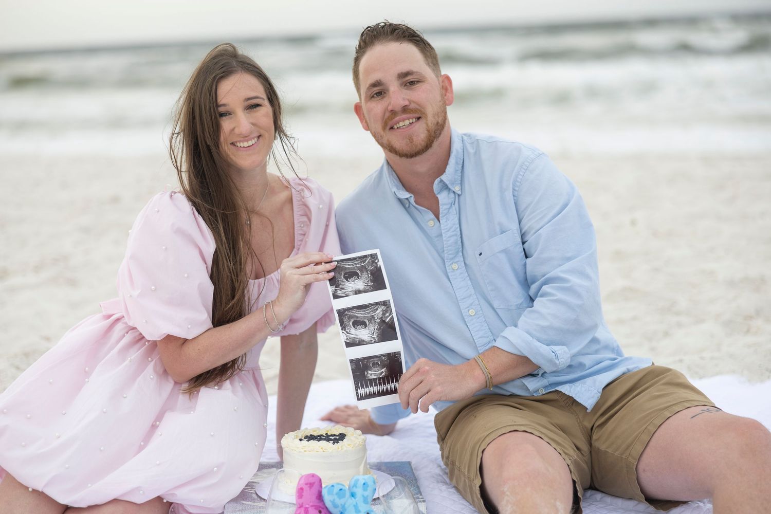 Couple sitting on beach blanket holding ultrasound photos for pregnancy announcement with ocean in background.