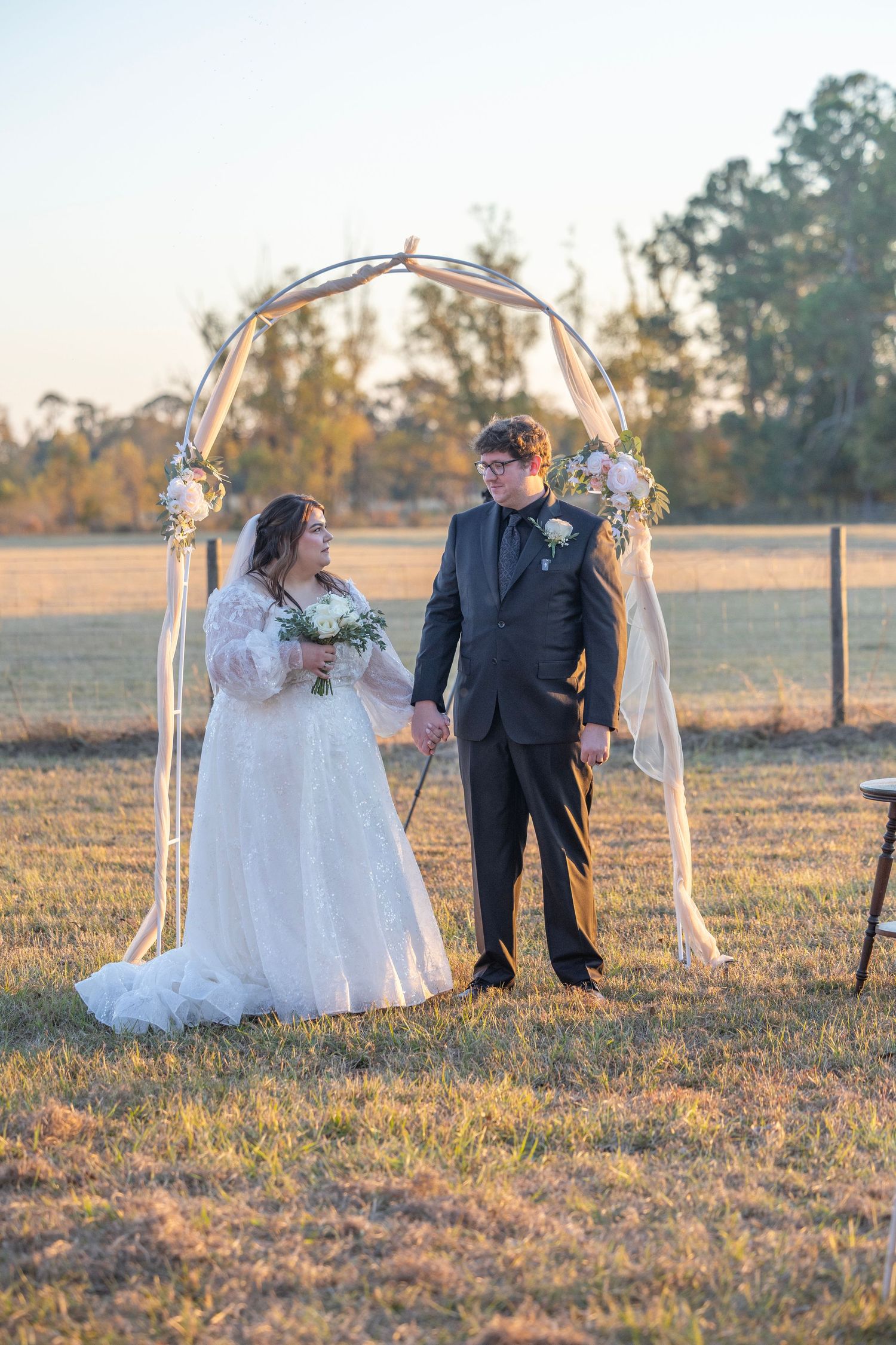 A couple in wedding attire shares a romantic moment at sunset in a rustic outdoor setting with draped fabric decor.
