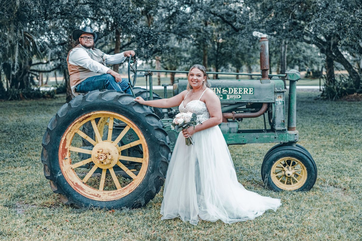A vintage tractor with yellow wheels serves as a rustic backdrop for a country wedding photo in an outdoor setting.