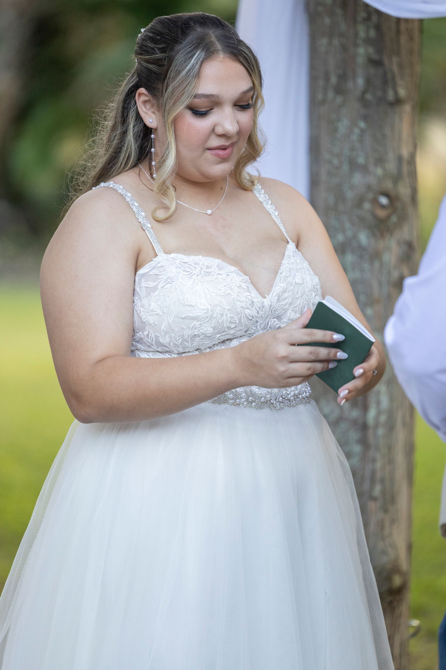 A bride in a white lace wedding dress holds a small book while standing outdoors near a tree.