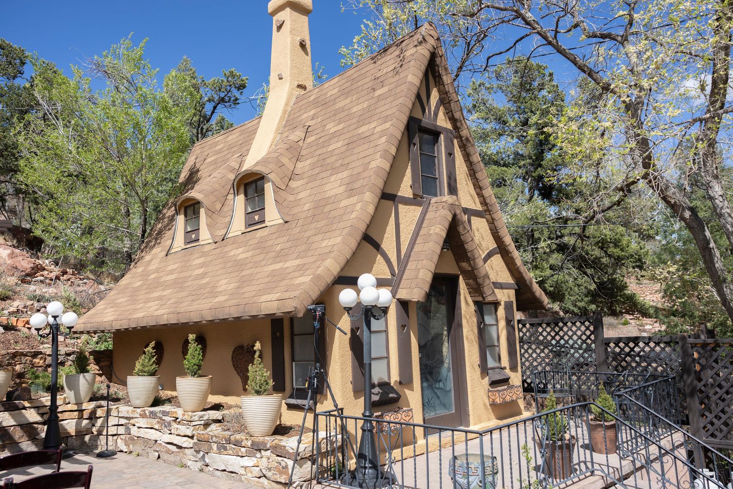 Whimsical storybook cottage with steep peaked roof and rustic wooden details stands among trees in a forest setting.