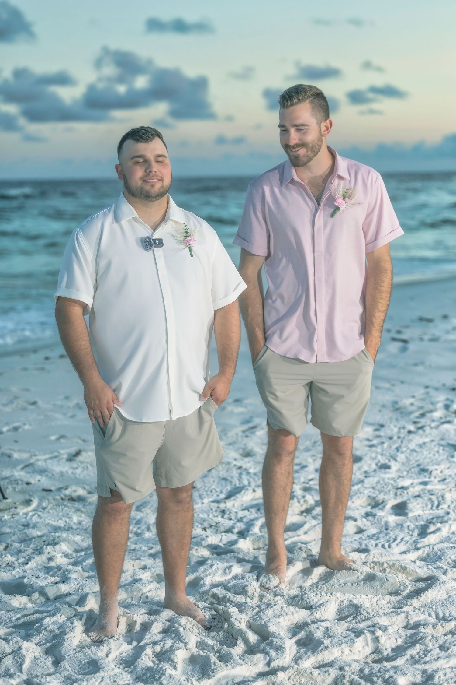Two people in coordinating white and pink outfits pose playfully on a pristine white sand beach at sunset.