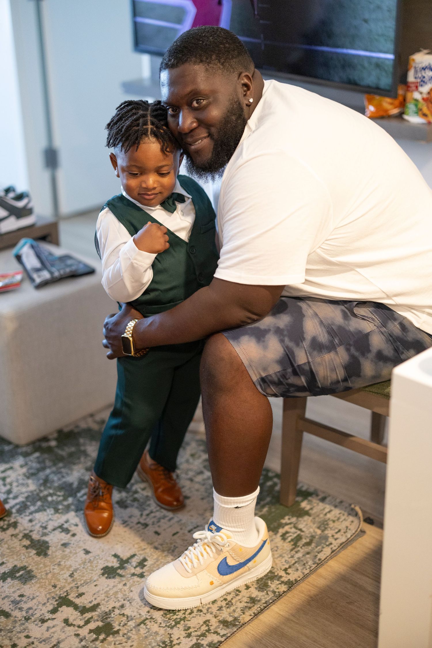 A person in a white shirt sits with a child in a green uniform on a patterned rug in a well-lit room.