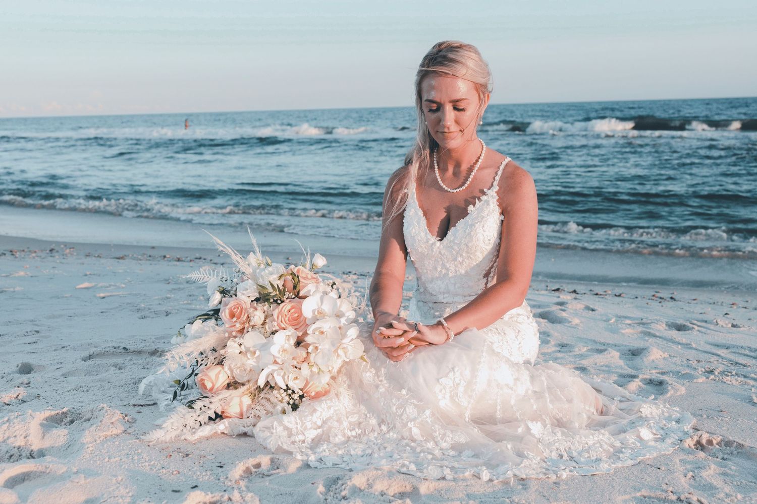 A bride in a white dress sits on a beach at sunset next to a beautiful floral arrangement in soft pink tones.