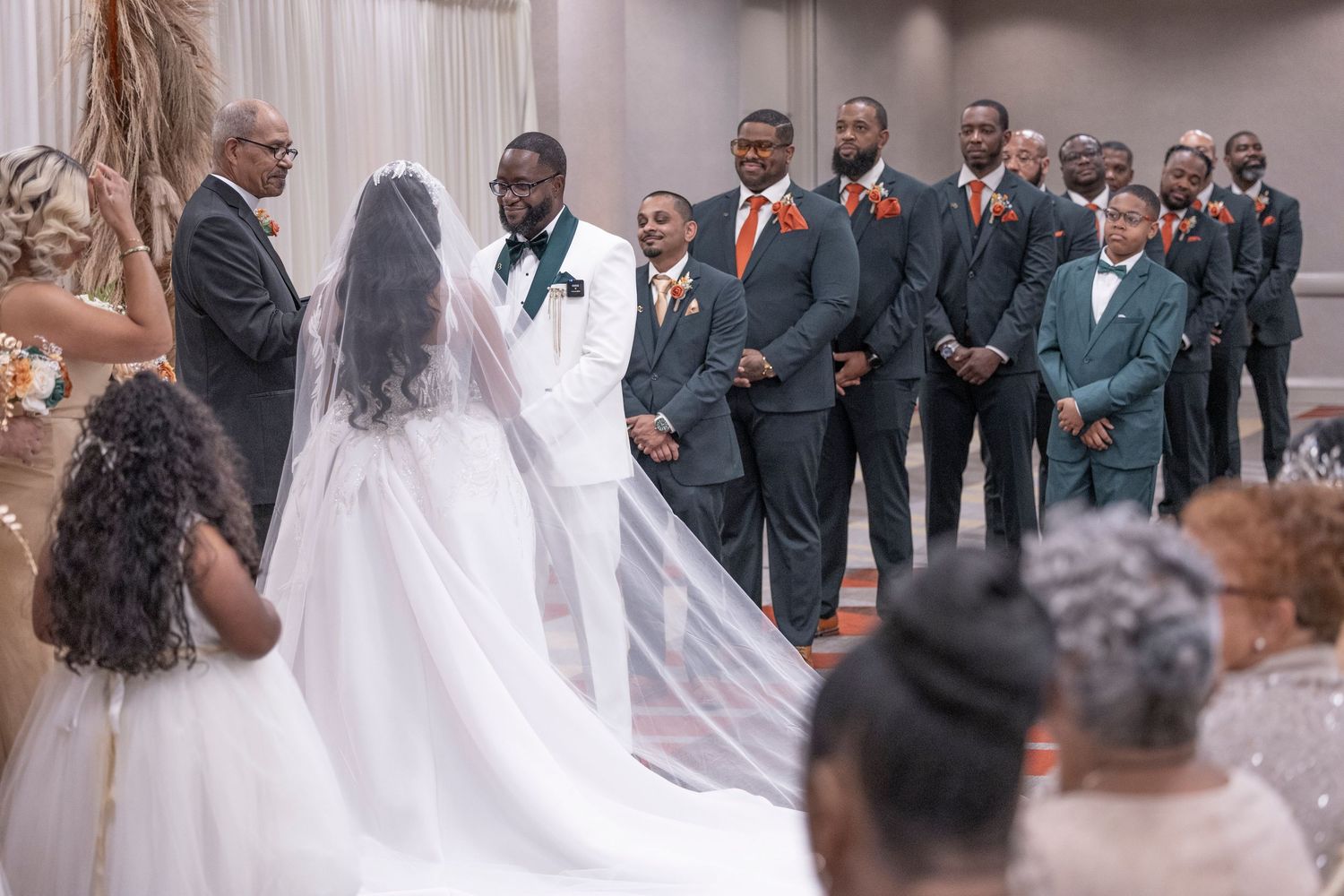 A wedding ceremony in progress with a bride in a flowing white veil and groomsmen lined up in charcoal gray suits.