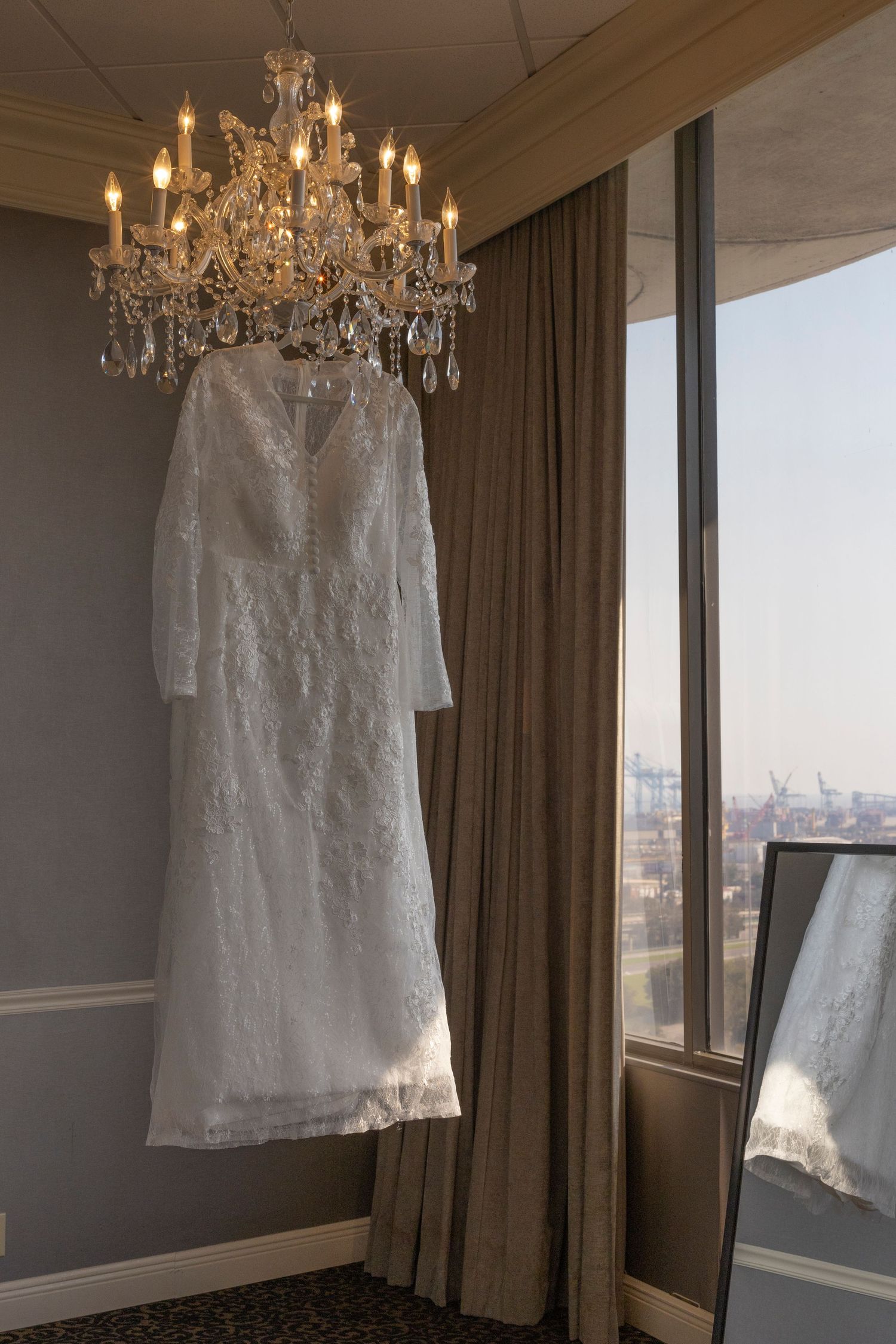 A white lace wedding dress hangs elegantly from a crystal chandelier in a room with city views through large windows.