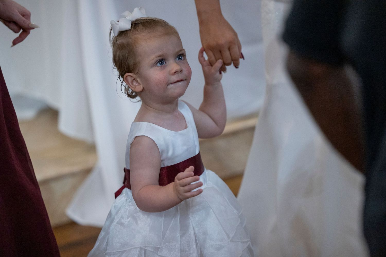 Young flower girl in white dress with burgundy sash holds hands with adults.