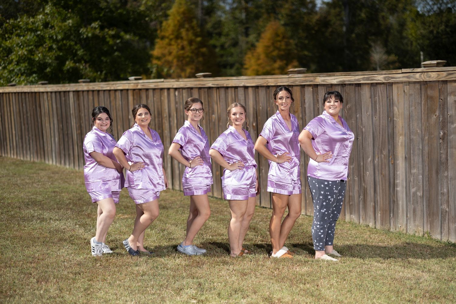 A group of bridesmaids in matching pink floral robes pose playfully in front of a wooden fence outdoors.