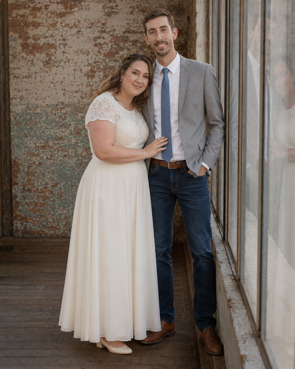 Couple poses together in wedding attire, woman in white lace dress, man in gray blazer and blue tie.