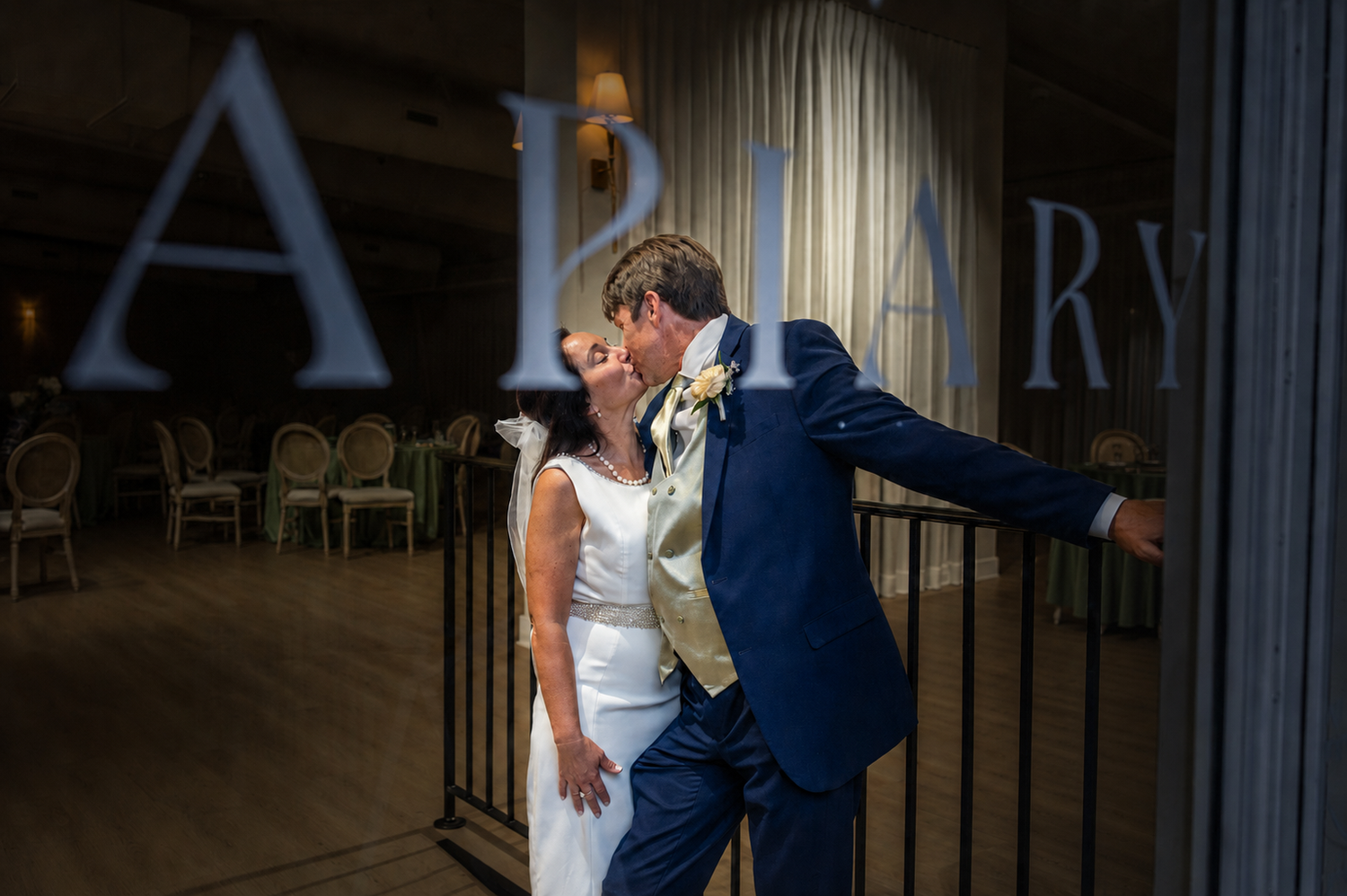 A bride and groom share a romantic kiss at their wedding reception venue, The Apiary, with elegant decor in the background.