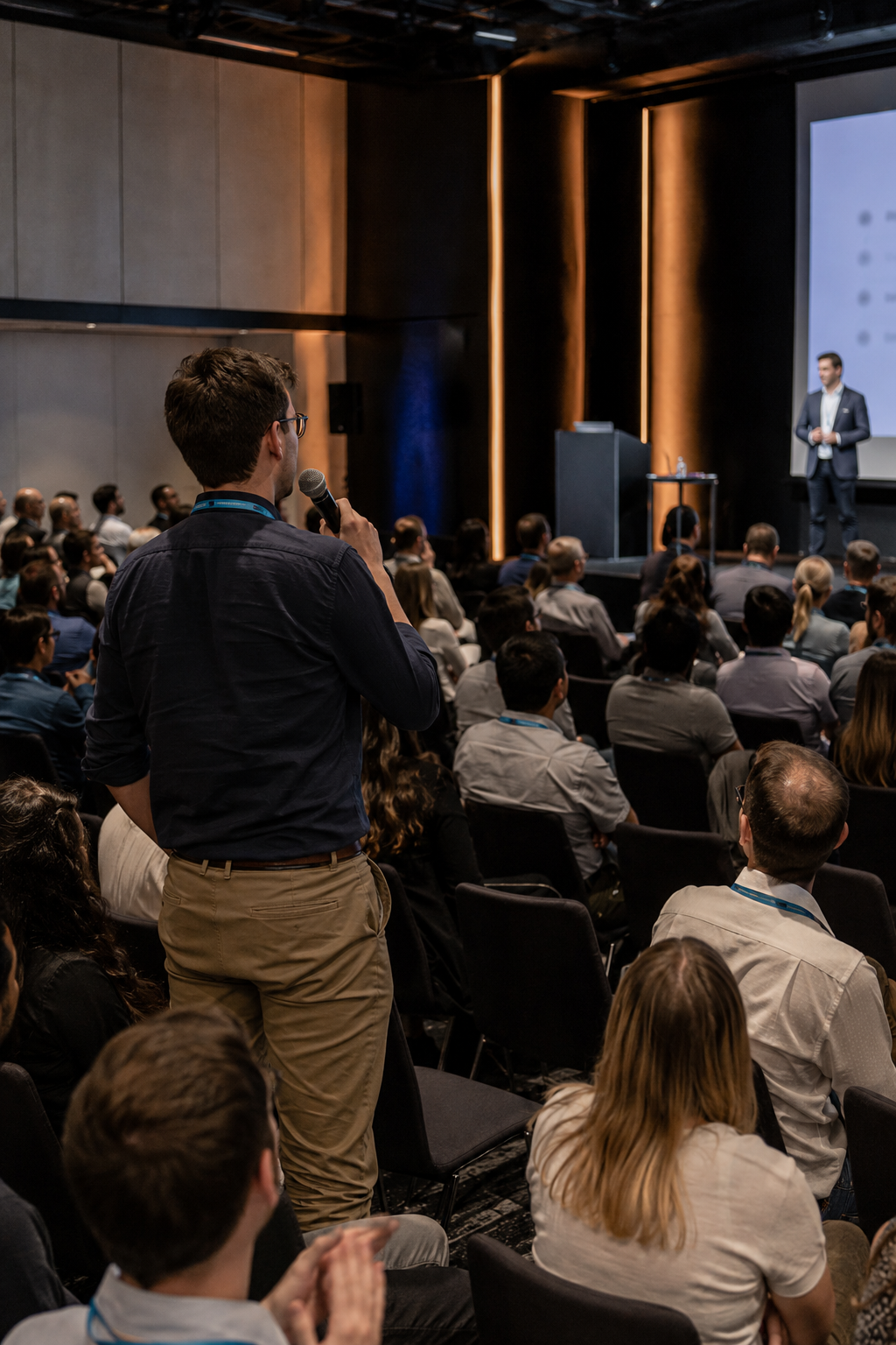 Audience member stands with microphone asking question at professional business conference presentation.