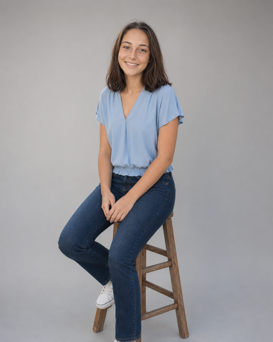 Young woman smiling, seated on wooden stool, wearing light blue top and dark jeans, white sneakers, gray background.