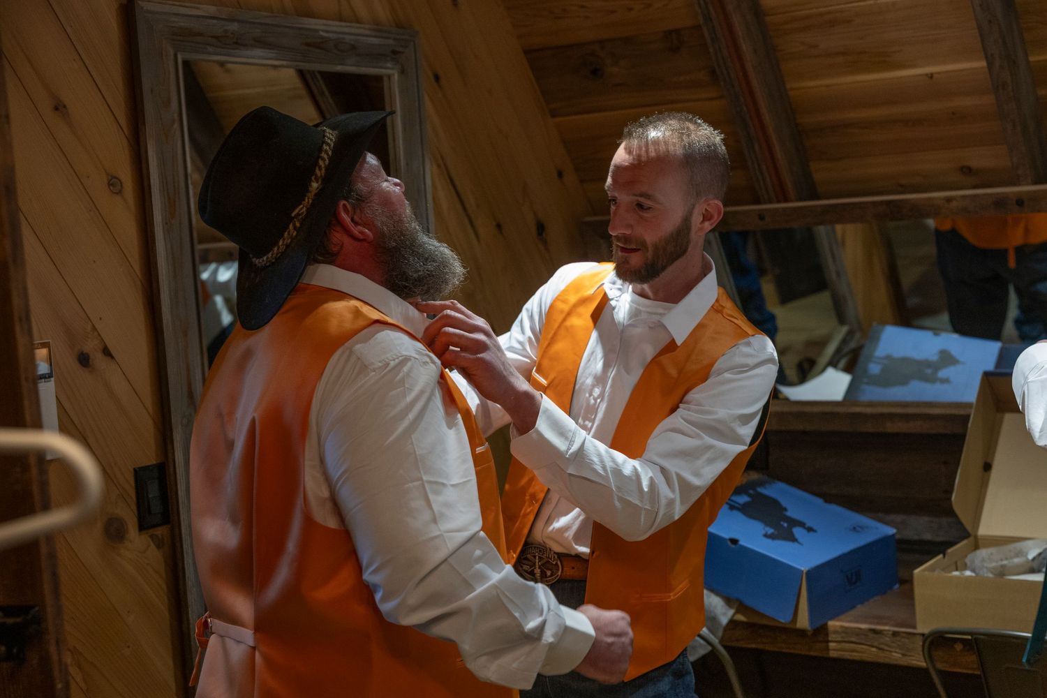 Two men in matching orange vests and white shirts help each other dress in a rustic wooden cabin interior.