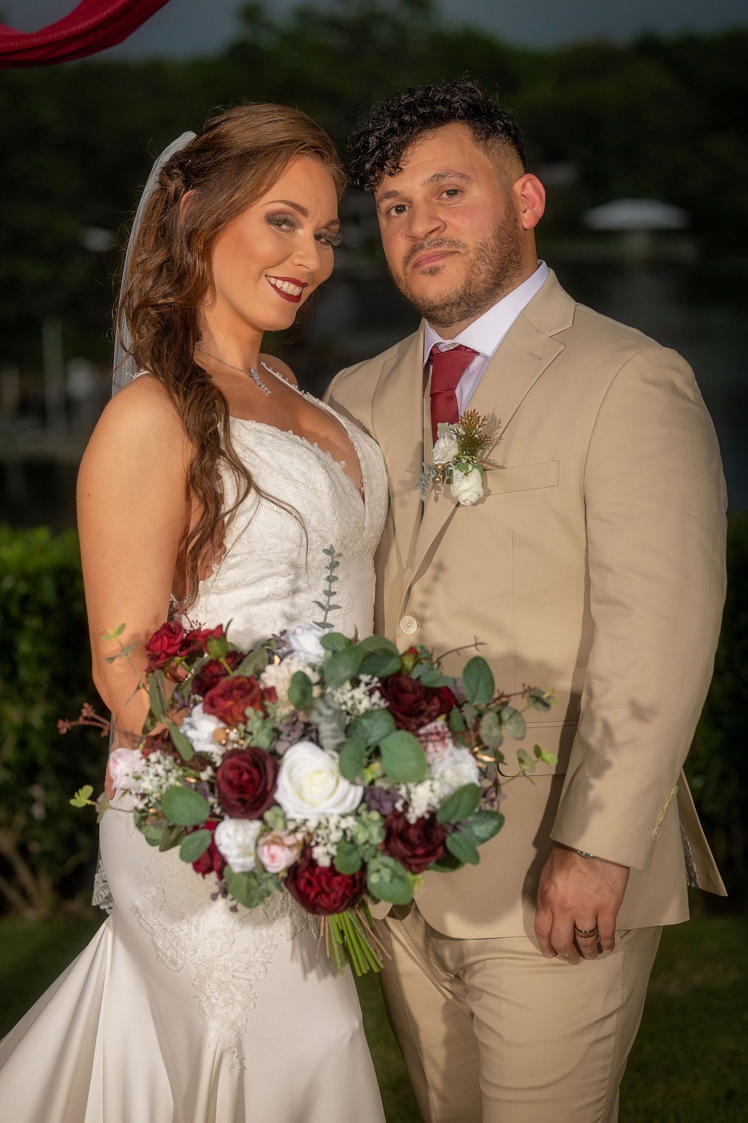 A bride in a white lace gown and groom in tan suit pose together with a red and white rose wedding bouquet at dusk.