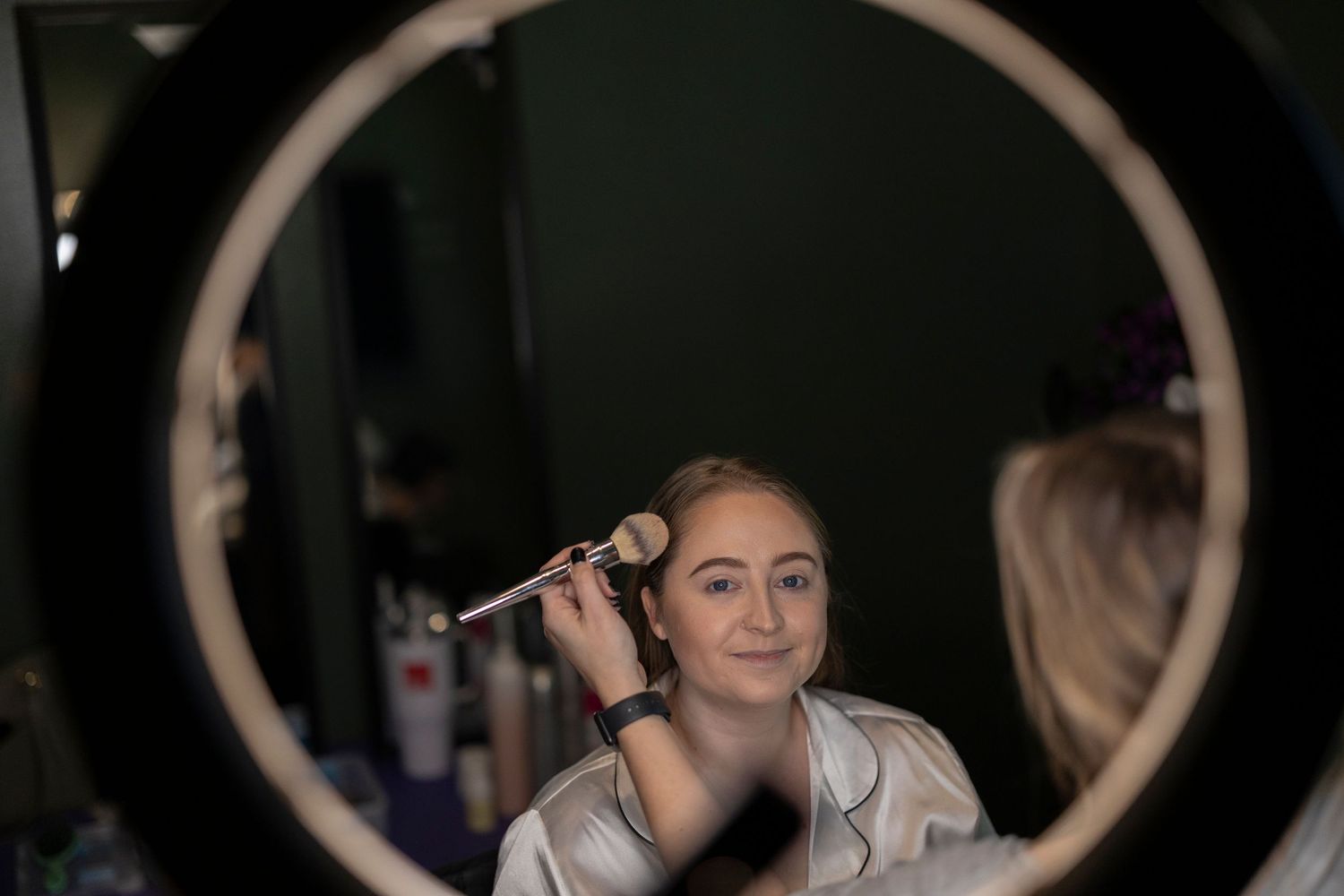 A reflection in a circular mirror shows a person styling hair in a dark room with professional equipment around.